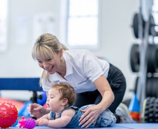 Adult assisting a baby on a blue mat with colorful toys around in a bright room.